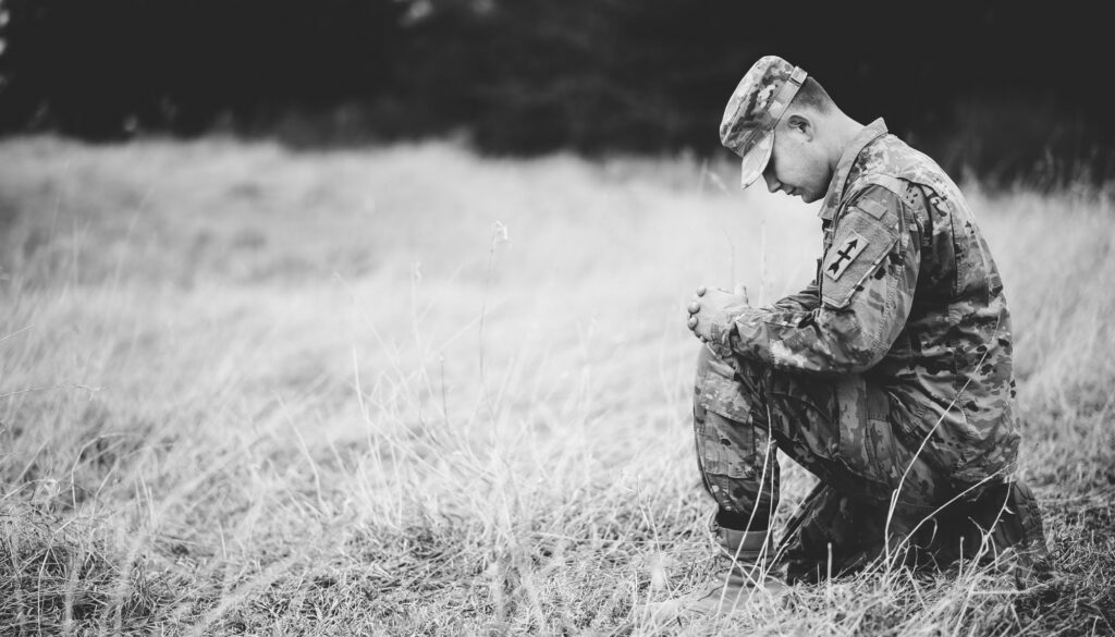 grayscale-shot-young-soldier-praying-while-kneeling-dry-grass-Web-Legacy